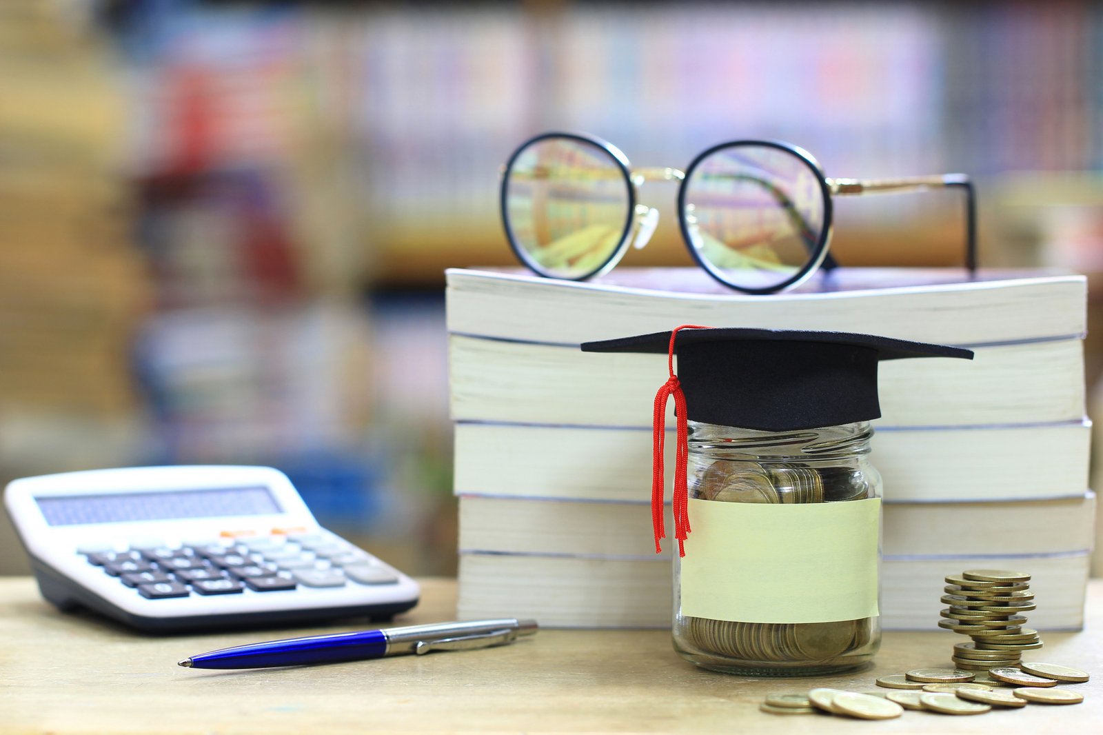 Graduation cap on a jar of coins beside a calculator and books
